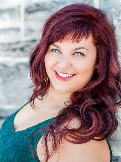 Photo of a person with long red hair, wearing a blue tank top, standing in front of a brick wall.