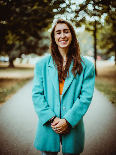 Person with long hair, wearing a teal blazer, smiling while standing in the middle of a wooded path.