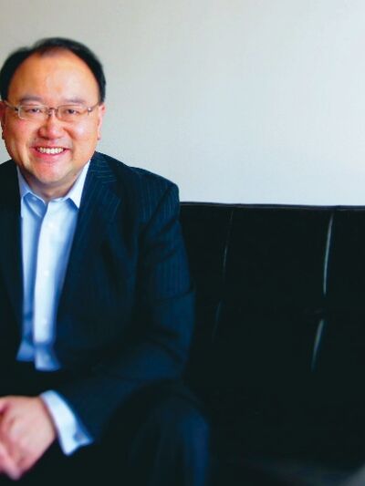 Andrew Kwan is sitting on a black chair, smiling.