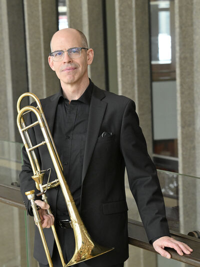 Photo of a man in a suit and glasses, holding a trombone, with windows in the background.
