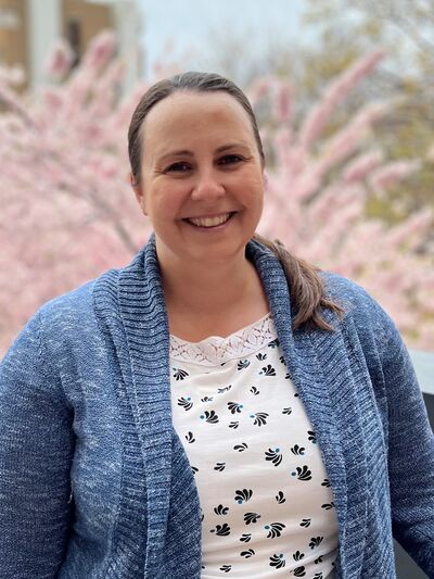 Person wearing a blue cardigan, smiling and posing in front of a cherry blossom tree in bloom