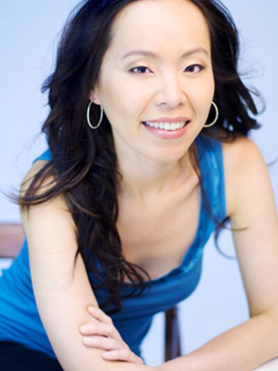 A headshot of a person with long black hair and hoop earrings, wearing a black tank top, leaning on a table.