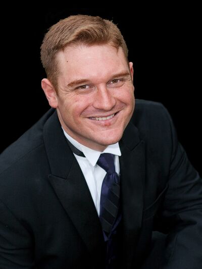 A headshot of a person wearing a suit, smiling and looking up at the camera, set against a black background.