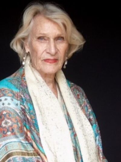 Headshot of a person wearing a colorful blouse and white pagmenia, looking at the camera with a confident expression against a dark background.