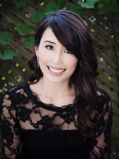 Person with long hair, wearing a black dress and smiling, standing in front of a lattice panel with leaves, creating a natural and elegant atmosphere.