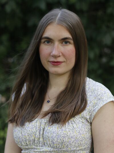 Young person with long hair wearing a white top. Blurred greenery in the background