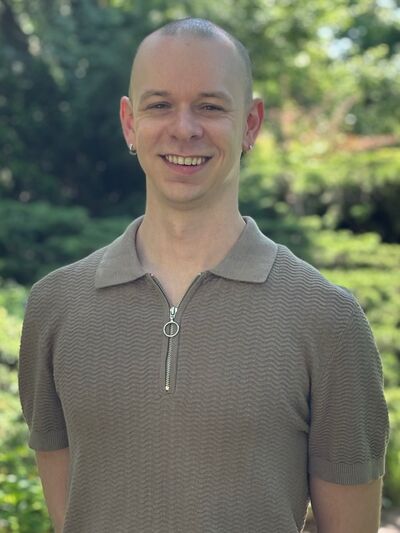 Person with buzz cut wearing in brown shirt. Blurred greenery in the background