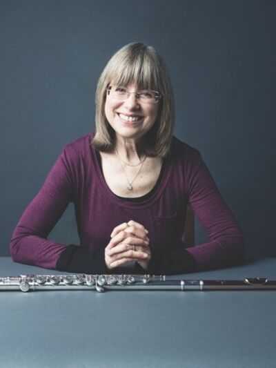Person wearing a purple shirt, sitting at a table with their hands interlaced and a flute placed in front of them, smiling warmly against a dark table and background.