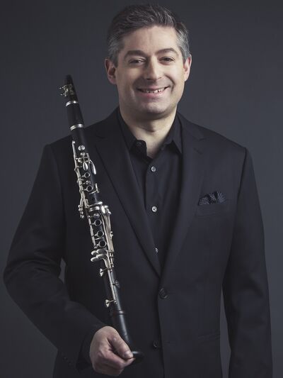 Person wearing a black suit, holding a clarinet and smiling, standing in front of a dark background, exuding sophistication and warmth.