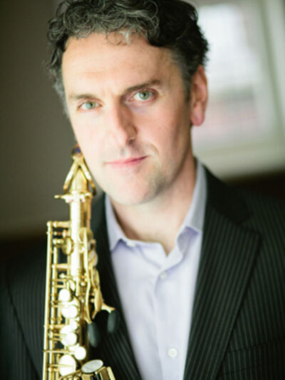 A headshot of a person wearing a white shirt and black suit jacket, holding a saxophone.