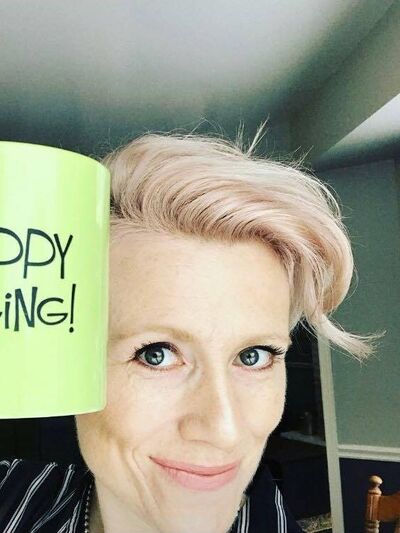 A person with short blonde hair grins while holding a green mug with the words 'happy singing' written on it, next to their face.
