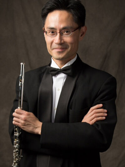 Person wearing a tuxedo, holding a flute in front of a brown background, presenting an elegant and refined appearance.