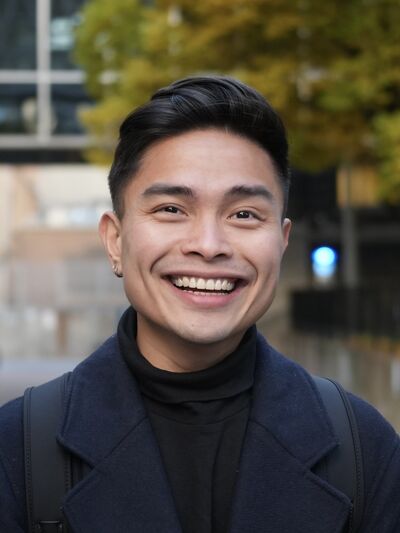 Headshot of a person wearing a black turtleneck and navy twill coat, smiling while standing in front of a tree and building.