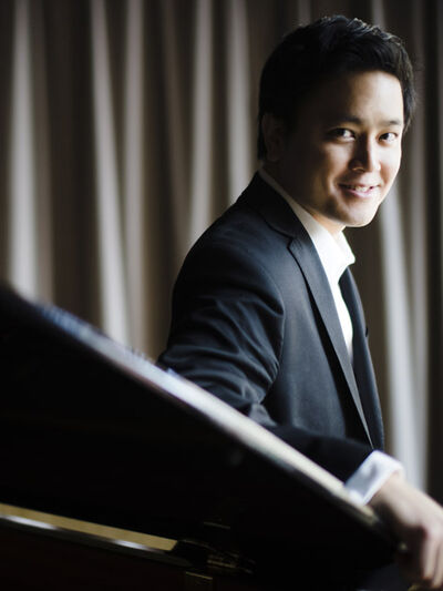 Person wearing a suit, leaning on a grand piano with curtains in the background, smiling.