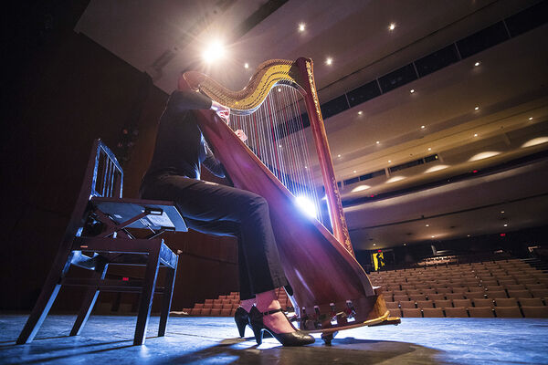 Person in performance attire playing the harp on stage, performing for an empty auditorium, with rows of seats and stage lighting in the background.