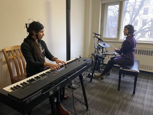 Two people wearing EEG caps, one playing a keyboard and the other playing an electronic drum set.