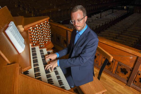 Man in a blue suit playing the organ in a church, focused on the keys while the large pipes of the organ rise behind him, set against the church’s architectural interior.