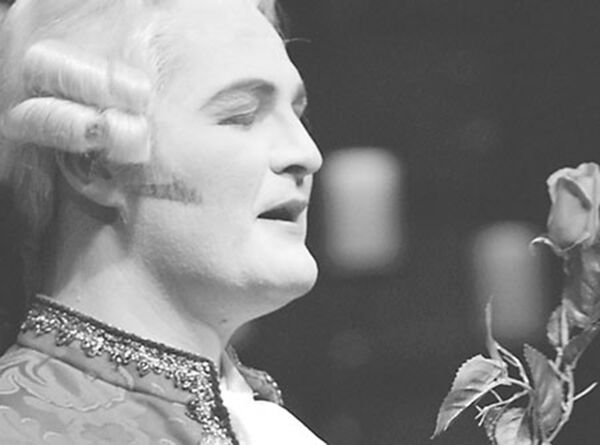 Black and white photo of a person in an Elizabethan costume and wig, singing to a rose.