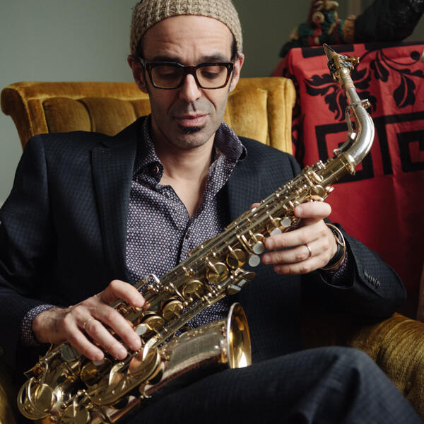 Michael Attias is sitting on a yellow chair with a saxophone in hand.