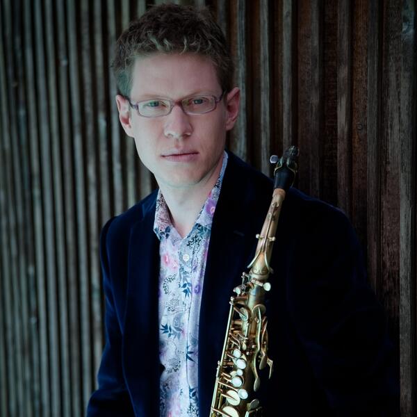 person standing in a blue blazer holding a saxophone in front of a wall