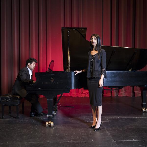 Vocalist and accompanist performing on stage, with the vocalist singing and the accompanist playing a grand piano, set against a red curtain backdrop.