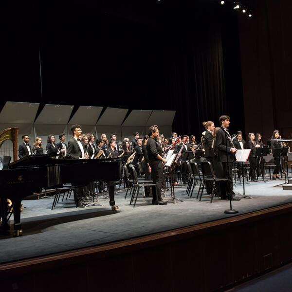 Large ensemble bowing to the audience on a larger stage, illuminated by stage lights, showing appreciation after their performance.