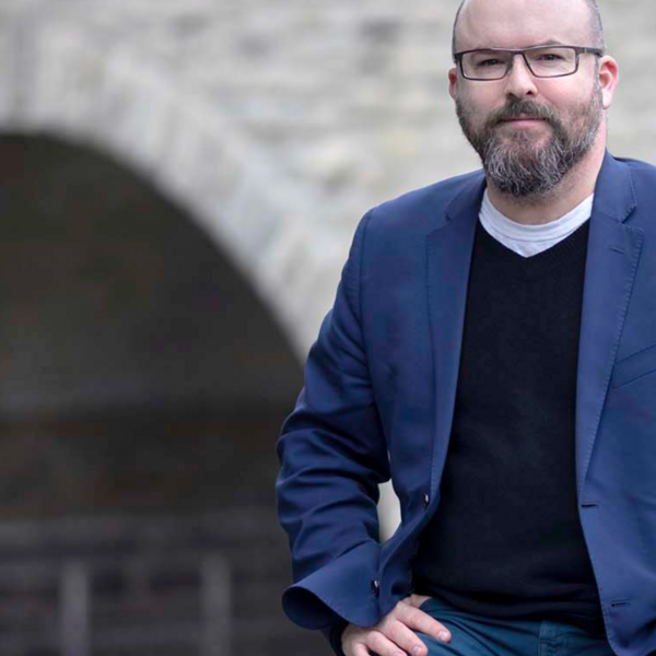 A person with glasses is wearing a black sweater, navy blazer, and jeans, posed in front of an old stone building.