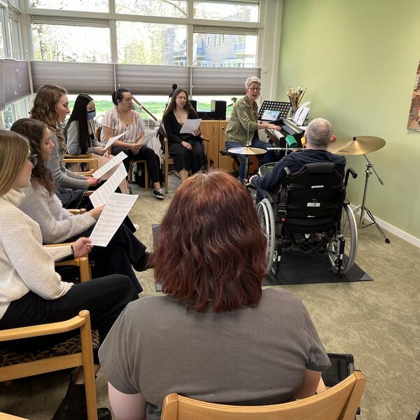 Man in a wheelchair and a piano accompanist facilitating a music class, with students engaged in the lesson.