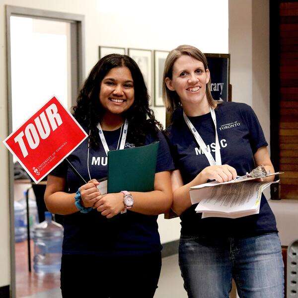 A Student Ambassador tour guide holds a Tour sign, and poses for a picture with a U of T Music staff member.